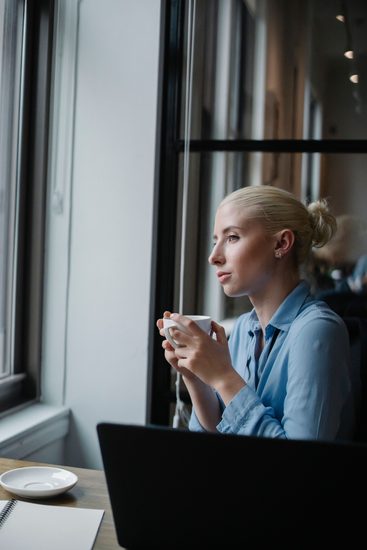 Frau hält eine Tasse Kaffee in der Hand und blickt nachdenklich aus dem Fenster und denkt 'arbeitslos geworden was tun'